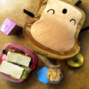K&ouml;nnte beinhalten: Eine Szene mit einem Lunchpaket mit einem affenf&ouml;rmigen Pl&uuml;schtier, einem Sandwich in einem roten Beh&auml;lter, einem Saftkarton, einem gr&uuml;nen Apfel und einem kleinen Beh&auml;lter mit M&uuml;sli. Die Artikel sind auf einer hellbraunen Oberfl&auml;che angeordnet.