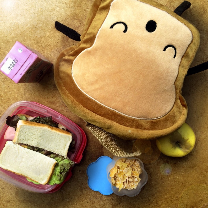 K&ouml;nnte beinhalten: Eine Szene mit einem Lunchpaket mit einem affenf&ouml;rmigen Pl&uuml;schtier, einem Sandwich in einem roten Beh&auml;lter, einem Saftkarton, einem gr&uuml;nen Apfel und einem kleinen Beh&auml;lter mit M&uuml;sli. Die Artikel sind auf einer hellbraunen Oberfl&auml;che angeordnet.
