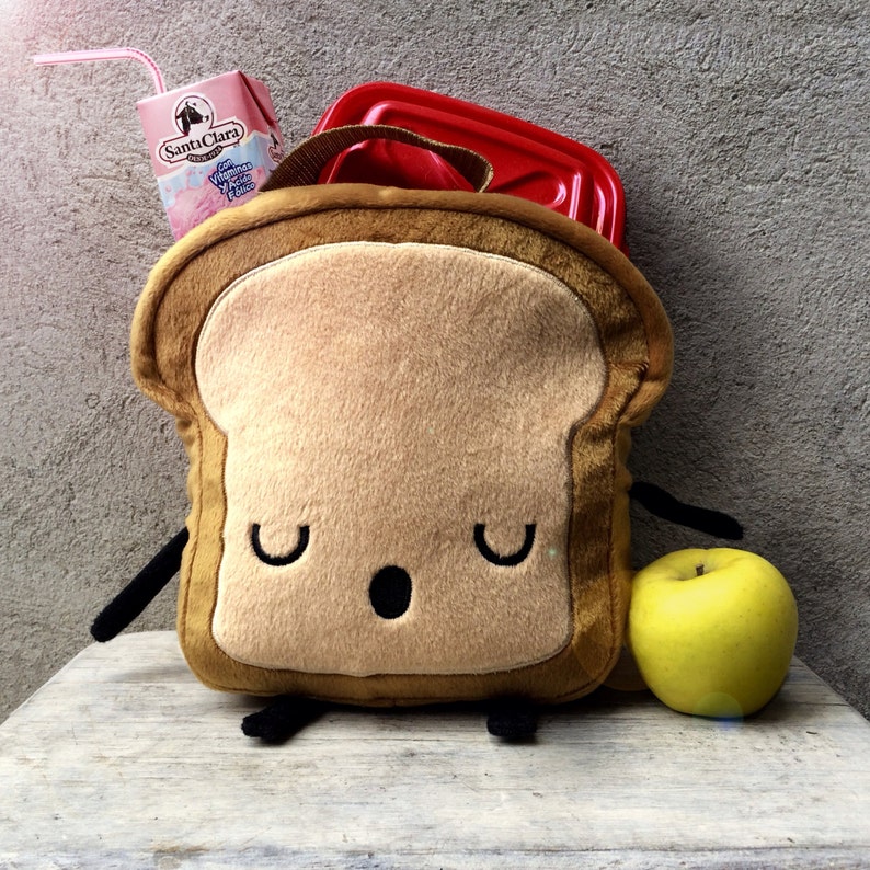 K&ouml;nnte beinhalten: Eine brotf&ouml;rmige Lunchtasche mit einem l&auml;chelnden Gesicht, schwarzen Armen und F&uuml;&szlig;en sowie einer roten Lunchbox im Inneren. Ein Karton Santa Clara Erdbeermilch und ein gelber Apfel liegen neben der Tasche. Die Tasche hat einen braunen Griff.