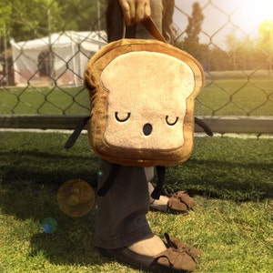 K&ouml;nnte beinhalten: Eine braune und beige Brotscheibe-Lunchtasche mit einem Cartoon-Gesicht mit geschlossenen Augen und einem kleinen Mund. Die Tasche hat kleine schwarze Arme und wird vor einem grasbewachsenen Hintergrund gehalten.