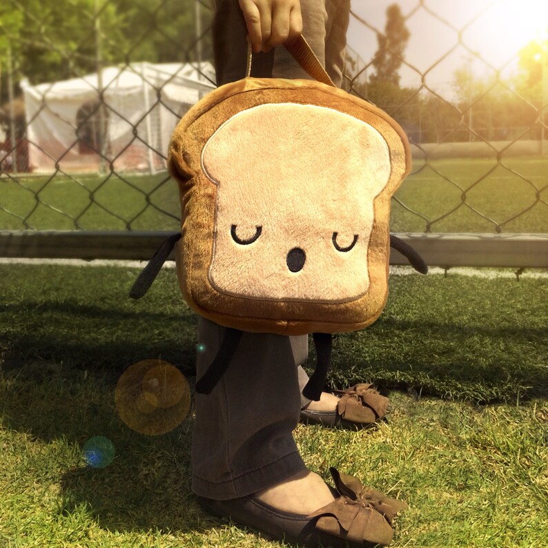 K&ouml;nnte beinhalten: Eine braune und beige Brotscheibe-Lunchtasche mit einem Cartoon-Gesicht mit geschlossenen Augen und einem kleinen Mund. Die Tasche hat kleine schwarze Arme und wird vor einem grasbewachsenen Hintergrund gehalten.