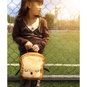 K&ouml;nnte beinhalten: Eine braune, pl&uuml;schige Brotscheiben-Lunchtasche mit einem l&auml;chelnden Gesicht und schwarzen Armen. Die Tasche wird vor einer Person gehalten, die sich in der N&auml;he eines Maschendrahtzauns und eines gr&uuml;nen Feldes befindet.