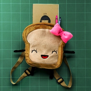 Op de afbeelding: Een grillige rugzak in de vorm van een toast, met een lachend gezicht en gesloten ogen. Hij heeft verstelbare bandjes, zwarte armen en een roze strik. Een klein notitieboekje en kleurpotloden zijn achter de rugzak gestoken.