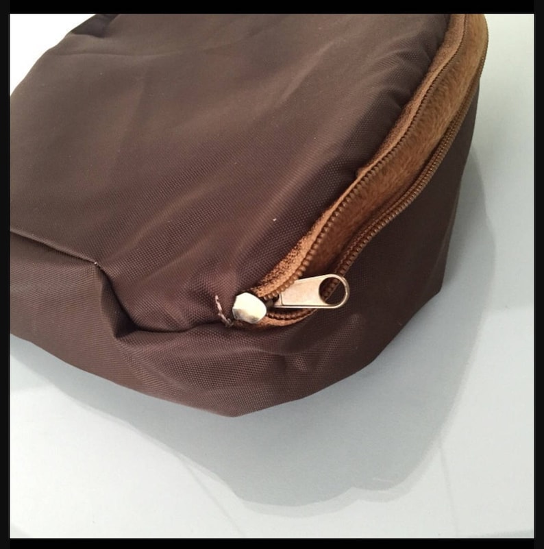 K&ouml;nnte beinhalten: Nahaufnahme einer braunen Stofftasche mit Rei&szlig;verschluss. Die Tasche hat eine rechteckige Form mit abgerundeten Kanten. Der Rei&szlig;verschluss ist goldfarben und verl&auml;uft entlang der Oberkante der Tasche.