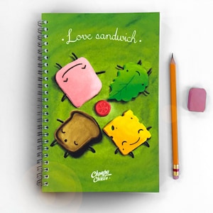 Puede incluir: Una libreta de espiral verde con una ilustración de dibujos animados de un sándwich. La ilustración presenta una pieza de carne rosa, una pieza de pan marrón, una hoja verde, una pieza de queso amarillo y un tomate rojo. El texto en la libreta dice "Love sandwich."