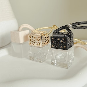 Może przedstawiać: Dwie małe szklane butelki z ozdobnymi metalowymi nakrętkami. Butelki wiszą na sznurku i są przeznaczone do użytku jako odświeżacze powietrza do samochodu. Jedna butelka ma złotą nakrętkę, a druga czarną.