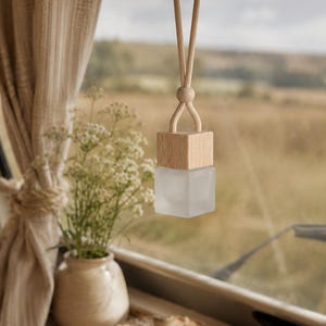 May include: A small, square, frosted glass bottle with a light-colored wooden cap, suspended by a beige cord with a wooden bead adjustment mechanism, hanging from a vehicle rearview mirror.