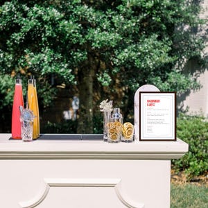 Puede incluir: Un carrito de bar blanco con una alfombra negra en la parte superior. Hay dos botellas de vidrio, una con un l&iacute;quido rosa y otra con un l&iacute;quido amarillo. Hay un vaso con una pajita de metal y una coctelera con rodajas de c&iacute;tricos secas. Un cartel enmarcado con el texto "DRINKS LIST" est&aacute; en el carrito de bar.