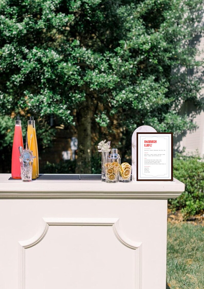 Puede incluir: Un carrito de bar blanco con una alfombra negra en la parte superior. Hay dos botellas de vidrio, una con un l&iacute;quido rosa y otra con un l&iacute;quido amarillo. Hay un vaso con una pajita de metal y una coctelera con rodajas de c&iacute;tricos secas. Un cartel enmarcado con el texto "DRINKS LIST" est&aacute; en el carrito de bar.