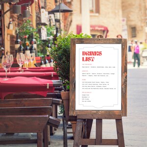 Puede incluir: Un cartel blanco con un t&iacute;tulo rojo y blanco "Drinks List" en un caballete de madera. El cartel enumera varias bebidas, incluyendo bebidas espirituosas, aperitivos, vinos, opciones no alcoh&oacute;licas y t&eacute; y caf&eacute;.
