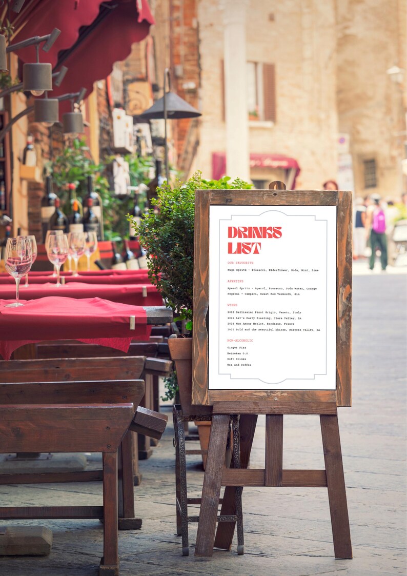 Puede incluir: Un cartel blanco con un t&iacute;tulo rojo y blanco "Drinks List" en un caballete de madera. El cartel enumera varias bebidas, incluyendo bebidas espirituosas, aperitivos, vinos, opciones no alcoh&oacute;licas y t&eacute; y caf&eacute;.