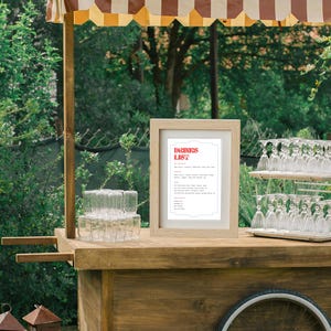 Puede incluir: Un carro de madera con un toldo a rayas, un letrero que dice "DRINKS LIST" y una variedad de cristaler&iacute;a. El carro est&aacute; al aire libre en un &aacute;rea cubierta de hierba.