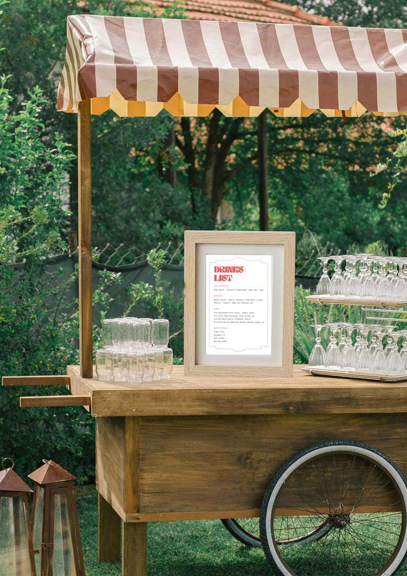 Puede incluir: Un carro de madera con un toldo a rayas, un letrero que dice "DRINKS LIST" y una variedad de cristaler&iacute;a. El carro est&aacute; al aire libre en un &aacute;rea cubierta de hierba.