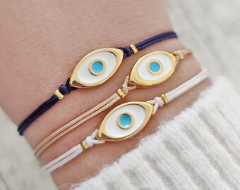May include: Four delicate bracelets with an evil eye design. Each bracelet features a gold-colored oval charm with a white enamel background and a blue center. The bracelets are strung on navy, beige, and white cords.