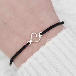 May include: A black string bracelet with a silver heart charm. The heart charm is open and has a delicate design.