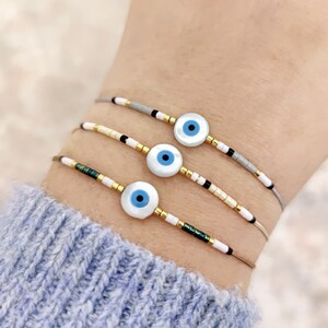 Könnte beinhalten: Drei übereinanderliegende Armbänder mit weißen und goldenen Perlen und einem blauen bösen Augen-Anhänger an jedem Armband.