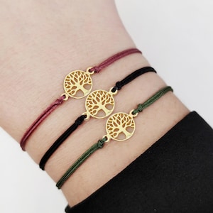 Puede incluir: Tres pulseras con un dije de árbol de la vida dorado. Las pulseras están en la muñeca de una persona. Las pulseras son burdeos, negras y verdes.