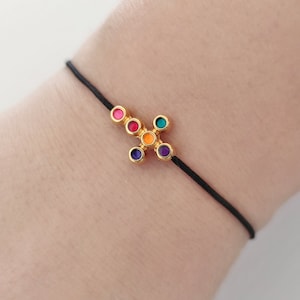 Peut inclure: Un bracelet en cordon noir avec un pendentif en forme de croix dorée. La croix est décorée de six cercles émaillés colorés en rose, orange, jaune, vert, violet et bleu.