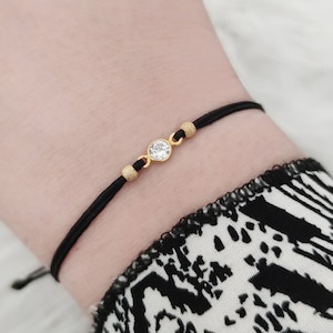 Puede incluir: Un brazalete ajustable de cuerda negra con un dije redondo dorado con una piedra de cristal transparente.