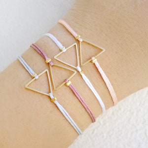 Puede incluir: Tres pulseras con un dije de triángulo dorado y cordones ajustables en blanco, rosa claro y rosa oscuro.