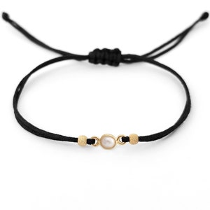 Puede incluir: Un brazalete de cuerda negra con una cuenta dorada a cada lado de una perla blanca.