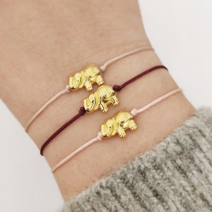 Può includere: Tre braccialetti d'oro con ciondolo a forma di elefante su un polso. I braccialetti sono realizzati con uno stringa sottile di colore bordeaux, crema e rosa chiaro.