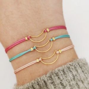 May include: Three gold crescent moon charm bracelets on a wrist. The bracelets are on pink, light blue, and light pink cords.