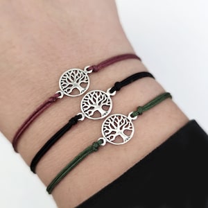 Puede incluir: Tres pulseras con un dije de árbol de la vida. Cada pulsera presenta un colgante circular plateado con un diseño de árbol. Los cordones son burdeos, negros y verde oliva. Las pulseras se llevan en una muñeca.