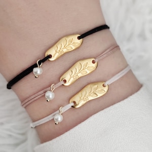 May include: Three gold bracelets with a leaf design and a small pearl charm on each. The bracelets are on a person's wrist and are made with black, pink, and white string.