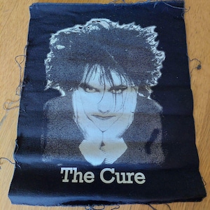 May include: Black fabric patch featuring a white image of Robert Smith, the lead singer of The Cure, and the text "The Cure" in white.