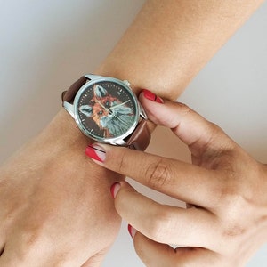 May include: A brown leather wristwatch with a silver face featuring a fox illustration. The watch is on a person's wrist with red nail polish.