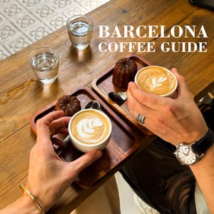 Op de afbeelding: Een houten tafel met twee kleine kopjes latte art, twee glazen water en twee gebakjes. De tekst "BARCELONA COFFEE GUIDE" wordt in witte letters weergegeven. De scène is van bovenaf vastgelegd.