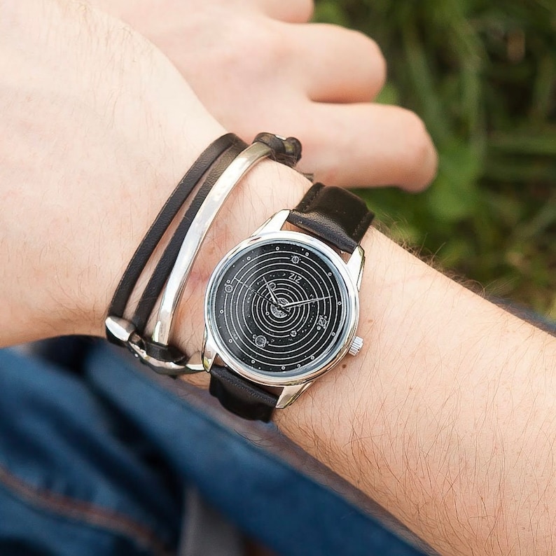 Pu&ograve; includere: Un orologio argentato con quadrante nero e cinturino in pelle. Il quadrante dell'orologio presenta un design di galassia con pianeti e stelle. L'orologio &egrave; indossato al polso.