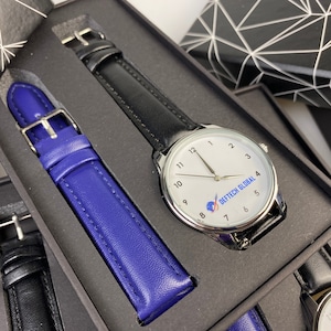 May include: A black leather watch with a white face and silver hands. The watch face has the text "DEFTECH GLOBAL" and a blue globe logo. The watch is in a black box with a blue leather watch band.