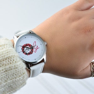 May include: White wristwatch with a silver band and a white face. The face has a red and green wreath with the word "Joy" written in red cursive.