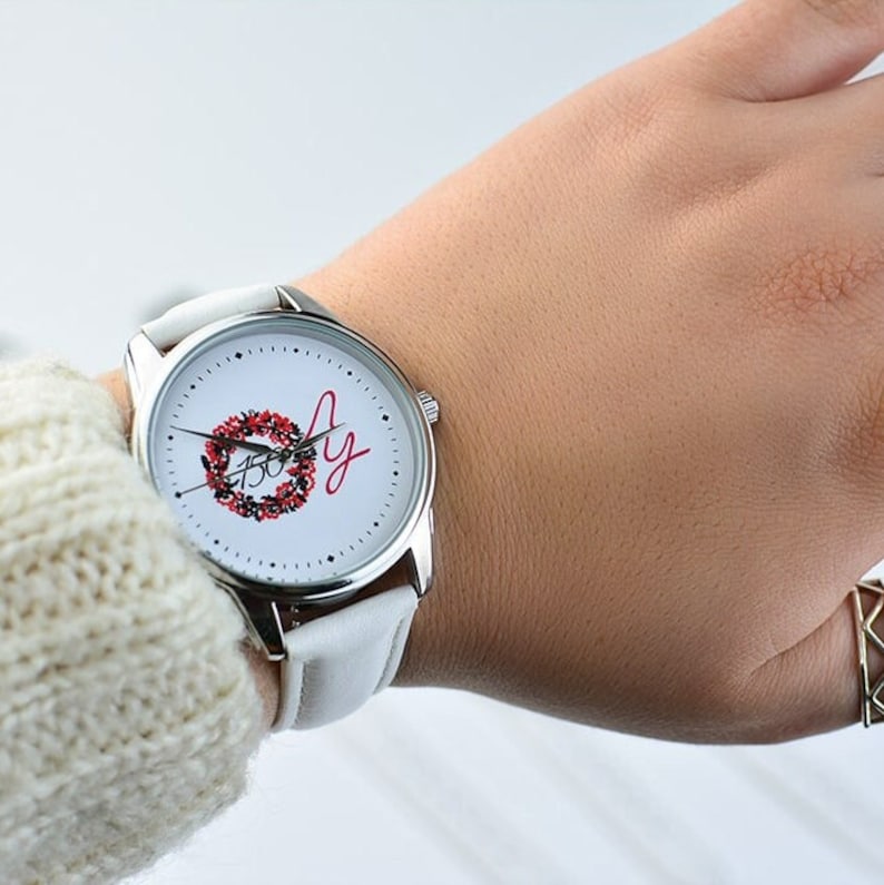 May include: White wristwatch with a silver band and a white face. The face has a red and green wreath with the word "Joy" written in red cursive.