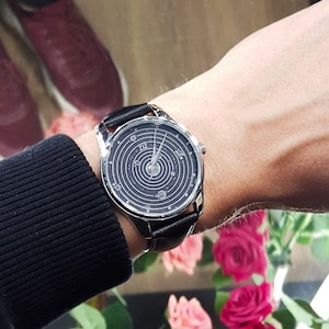 Pu&ograve; includere: Un orologio da polso in pelle nera con quadrante argentato e un design a sistema solare. Il quadrante dell'orologio ha la scritta "Z1Z" stampata su di esso.