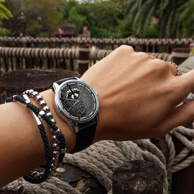 May include: A silver wristwatch with a black face and a black band. The watch face has a graphic of a raccoon with a cracked glass effect. The watch is worn on a wrist with a black and silver beaded bracelet.
