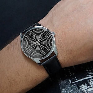 Pu&ograve; includere: Un orologio da polso argentato con un cinturino in pelle nera. Il quadrante dell'orologio ha uno sfondo nero con un design bianco di un sistema solare, che mostra pianeti e stelle.