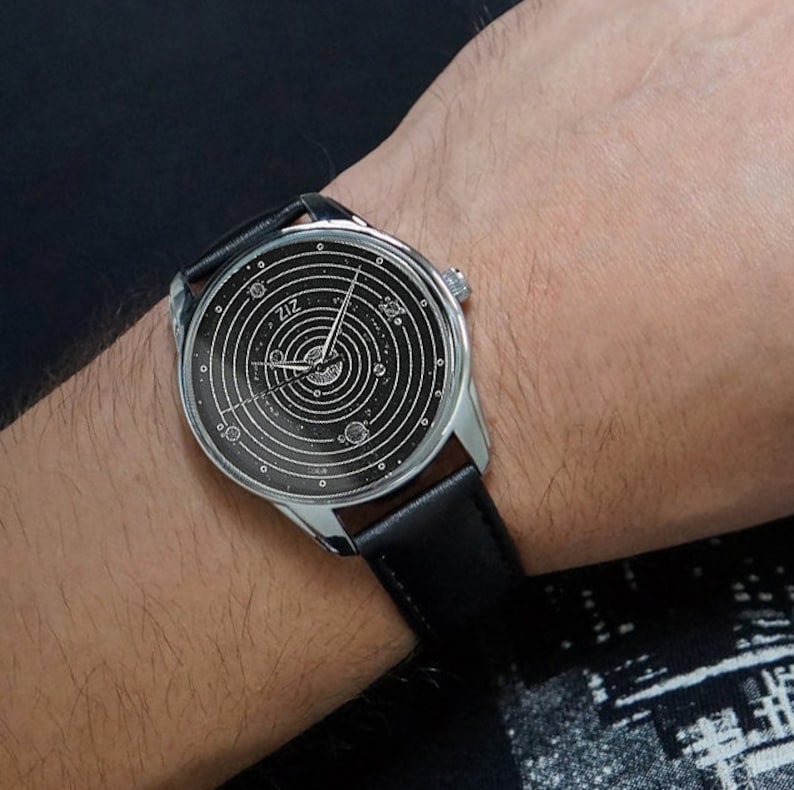 Pu&ograve; includere: Un orologio da polso argentato con un cinturino in pelle nera. Il quadrante dell'orologio ha uno sfondo nero con un design bianco di un sistema solare, che mostra pianeti e stelle.