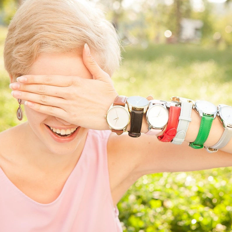 May include: A woman wearing a pink shirt and a stack of seven wristwatches with different colored bands. The watches have white faces and silver hands.