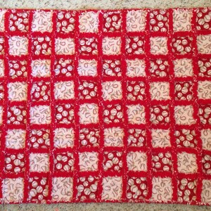 May include: A red and white rag quilt with a baseball theme. The quilt features a grid pattern of alternating squares. Some squares have a baseball print, while others have a horseshoe print. The quilt is made of soft fabric.