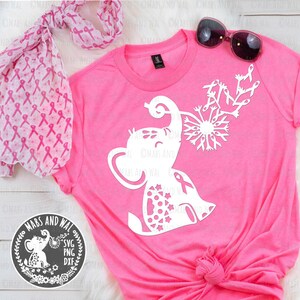 May include: A pink t-shirt with a white silhouette of an elephant blowing a dandelion made of pink ribbons. The elephant is wearing a pink ribbon. The t-shirt is on a white surface with a pink scarf and sunglasses.