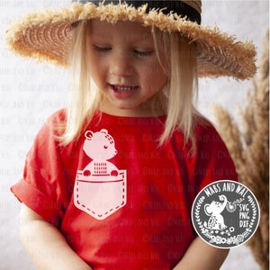 May include: A red t-shirt with a white pocket featuring a cartoon bear peeking out. The bear is wearing a sweater. The shirt is being worn by a child wearing a straw hat. The image also includes a logo with the text "MABS AND WAL" and the file types "SVG, PNG, DXF".