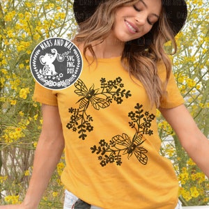 Op de afbeelding: Een geel T-shirt met een zwart bloemenontwerp met twee bijen. Het ontwerp is een cirkel met bijen en bloemen aan elke kant. De tekst "MABS AND WAL SVG PNG DXF" staat op een zwarte cirkel met een witte olifant erin.