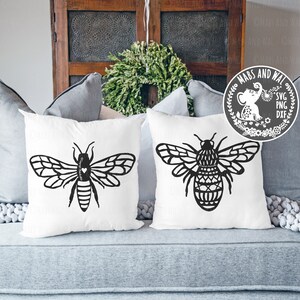 May include: Two white throw pillows with black bee designs. The bee on the left pillow has a heart on its body. The bee on the right pillow has a geometric pattern on its body. The pillows are on a gray couch.