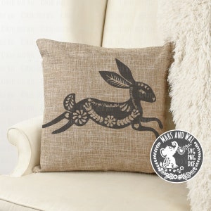 May include: A brown throw pillow with a black silhouette of a stylized rabbit with floral details. The pillow is on a white chair with a white fluffy blanket. The image also includes a black and white logo with the text "Mabs and Wal" and a small elephant illustration.
