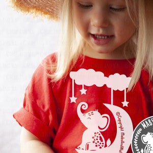 May include: A red t-shirt with a white design of an elephant sitting on a crescent moon with stars. The design includes the text "Copyright MabsAndWal".