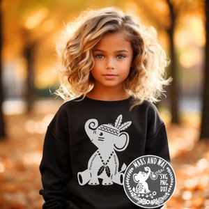 Puede incluir: Una niña pequeña con una sudadera negra con un gráfico blanco de un elefante con un tocado. El elefante sostiene plumas y cuentas. La sudadera tiene el texto "Happy Birthday". La imagen también incluye un gráfico circular con el texto "Mabs and Wal" y "SVG PNG DXF".