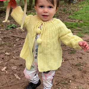 May include: A young child wearing a yellow crocheted cardigan with a white pom-pom detail, standing in a dirt yard.